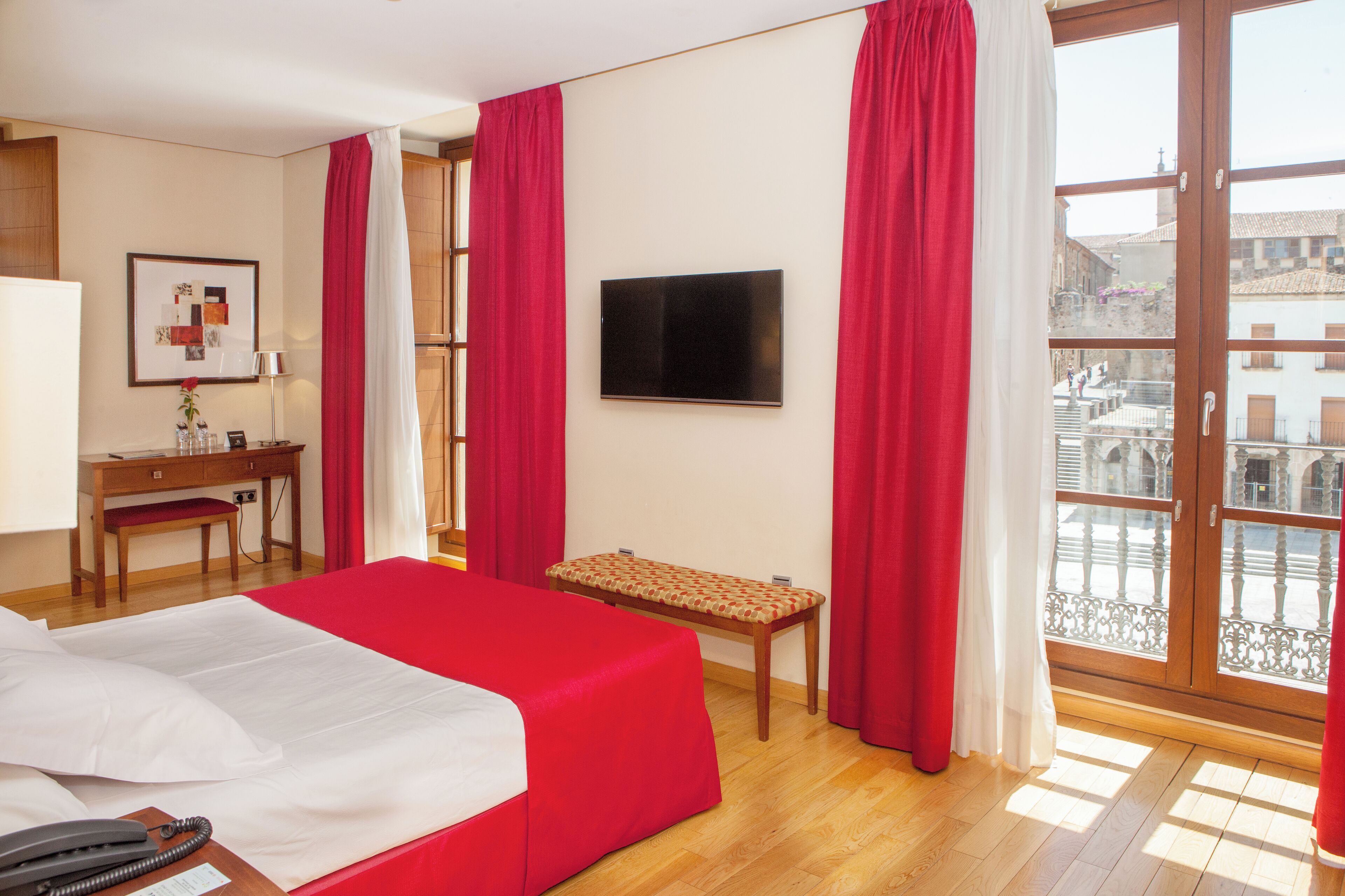 Habitación de hotel con cama, escritorio, silla, televisor y balcón con vistas a edificios.