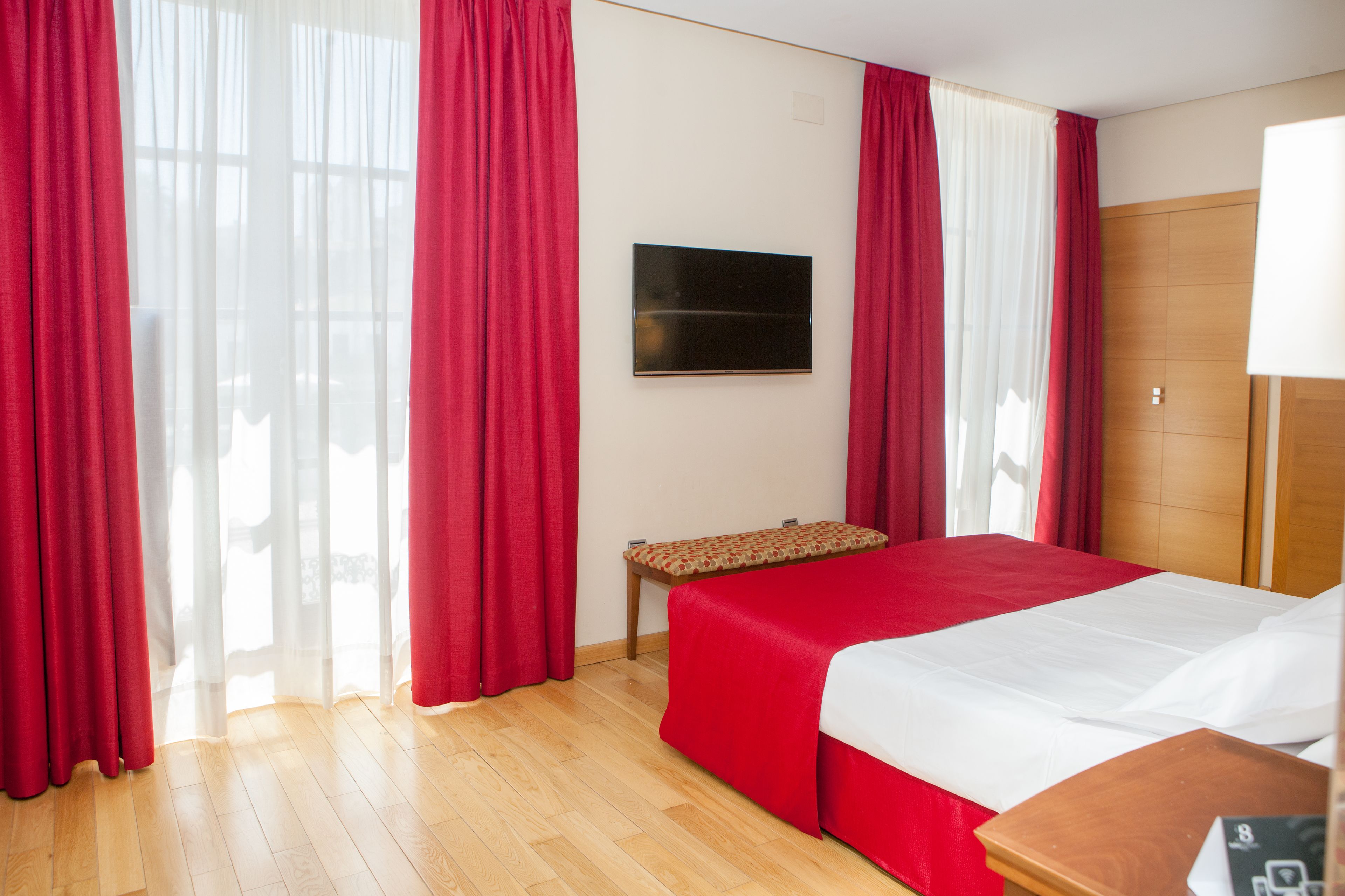 Een hotelkamer met een bed, een bankje, een televisie en rode gordijnen.
