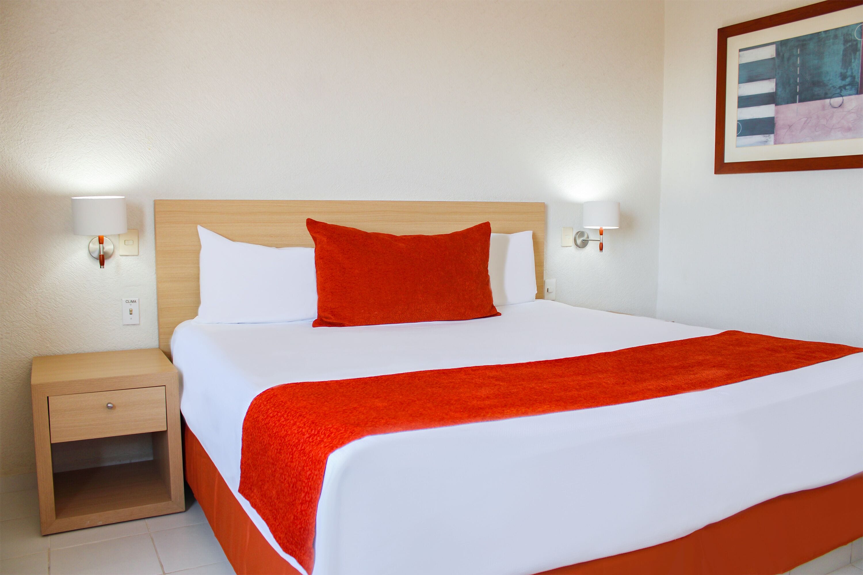 A neatly made bed with a red blanket and pillows, a wooden nightstand with a drawer, and two wall-mounted lamps.