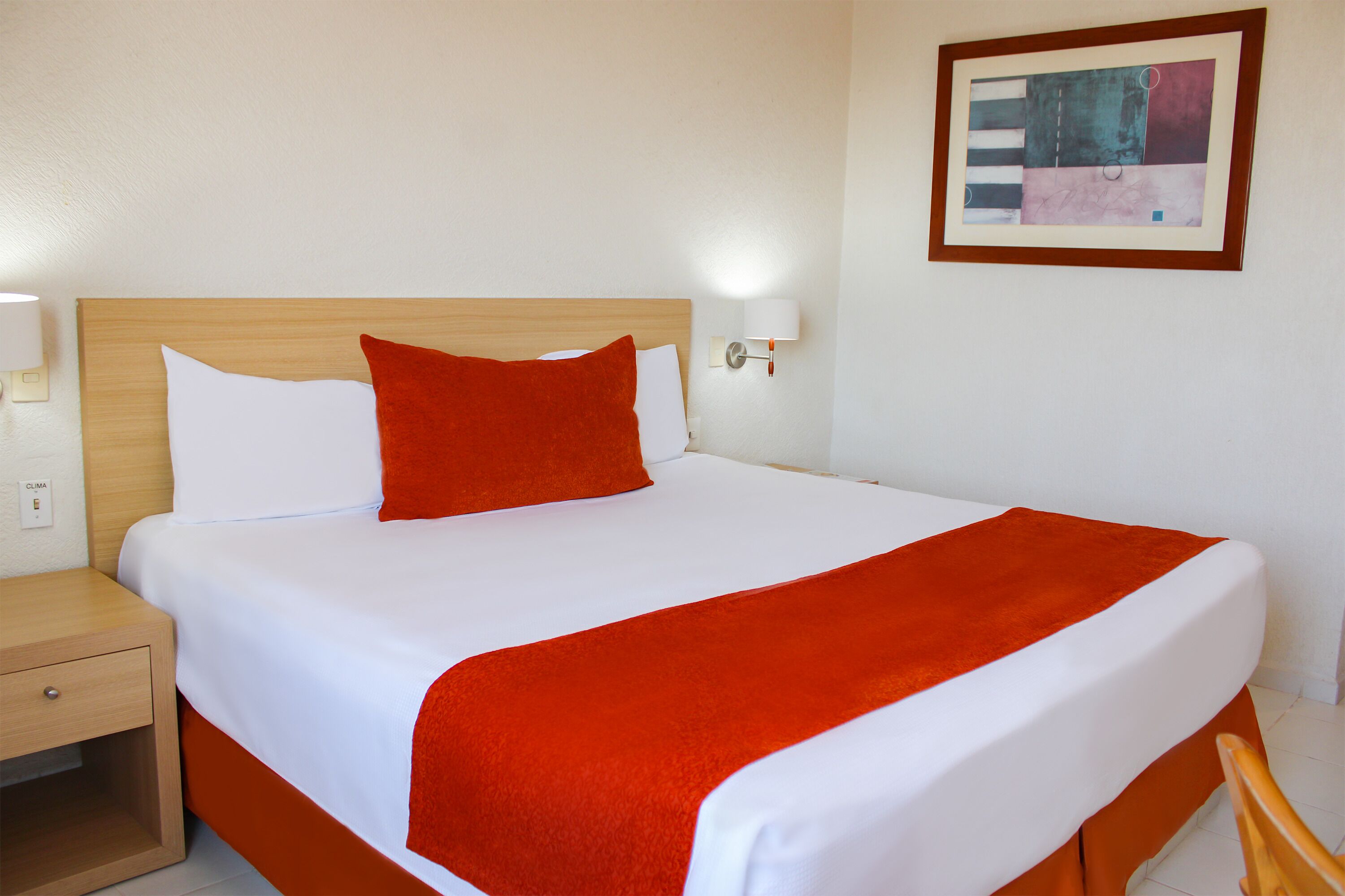 A neatly made bed with white linens and a red throw blanket, a wooden nightstand with a drawer, and a wall-mounted lamp.