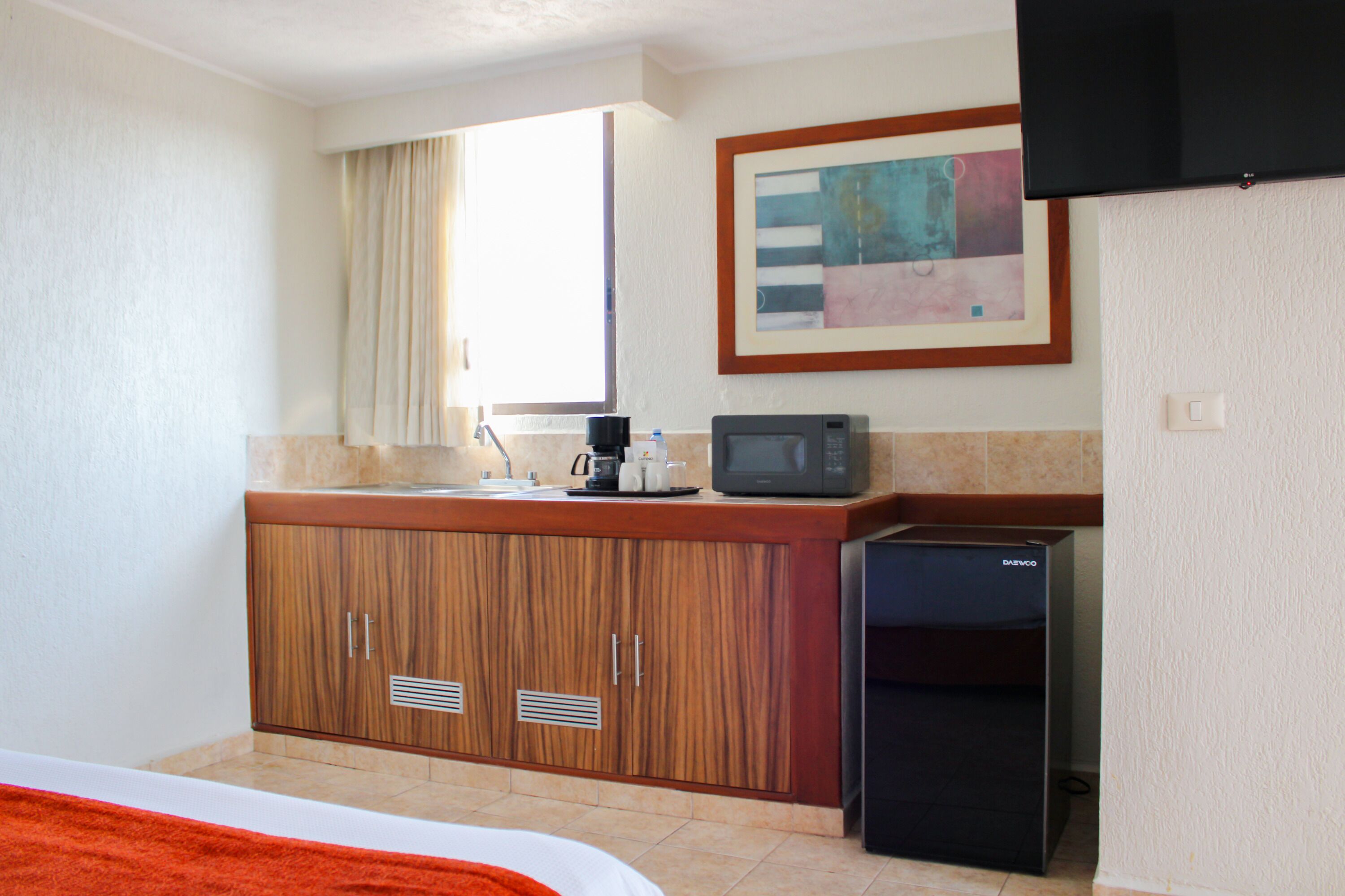 A compact hotel room with a kitchenette, a bed with an orange blanket, a flat-screen TV mounted on the wall, and a window with curtains.