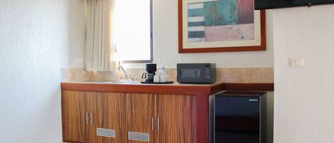 A compact hotel room with a kitchenette, a bed with an orange blanket, a flat-screen TV mounted on the wall, and a window with curtains.