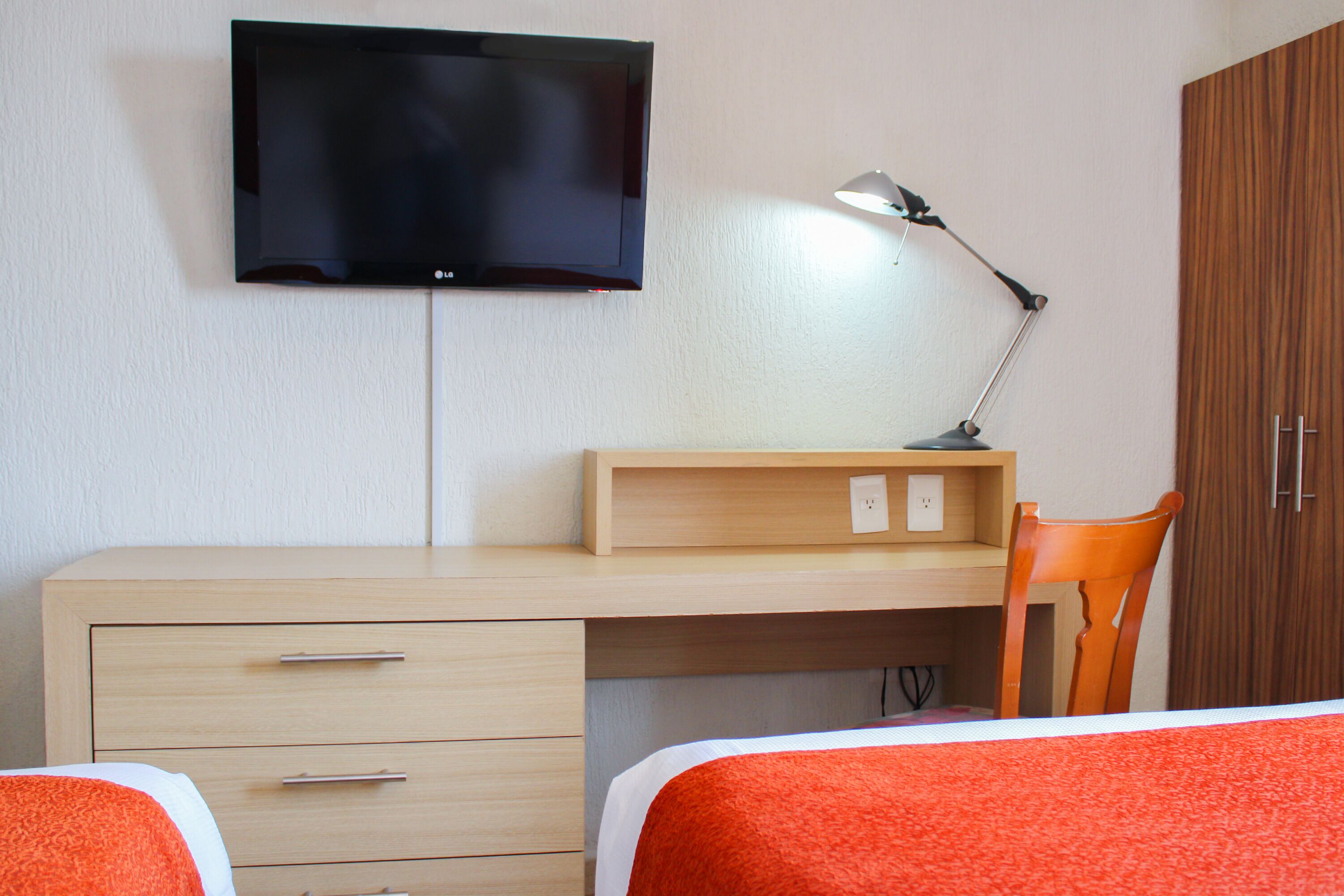 Une chambre à coucher avec un lit, un bureau et une chaise, un téléviseur fixé au mur et une lampe.