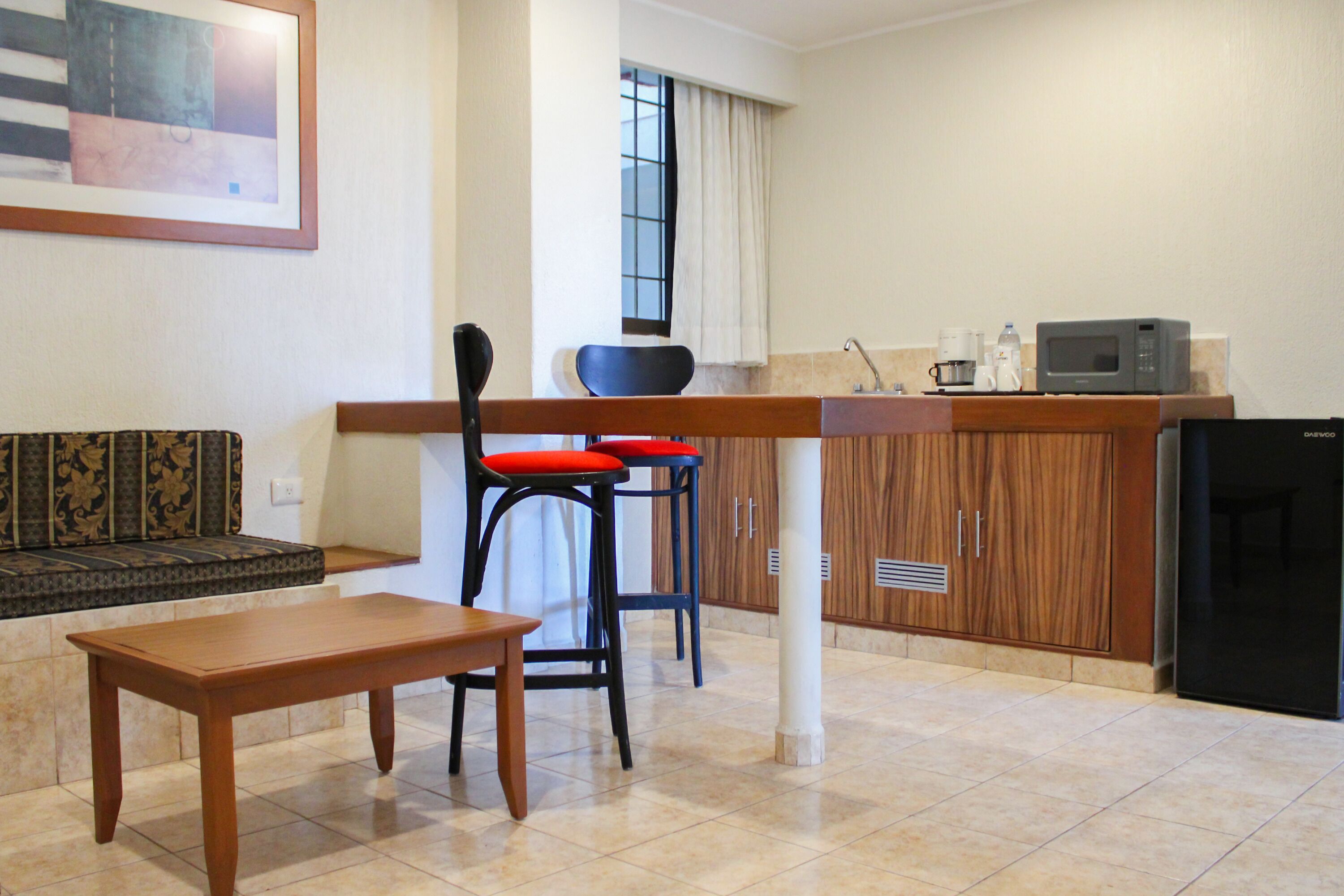 Un espace de vie compact comprenant une table à manger en bois, deux tabourets de bar, un canapé et un coin cuisine équipé d’un four à micro-ondes et d’un réfrigérateur.