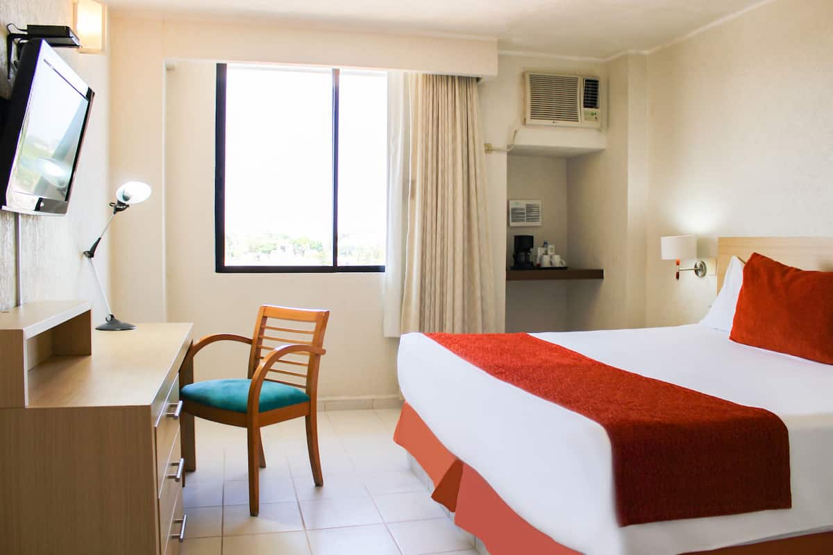 Une chambre d’hôtel avec un lit, un bureau et une chaise, une télévision et une fenêtre avec des rideaux.