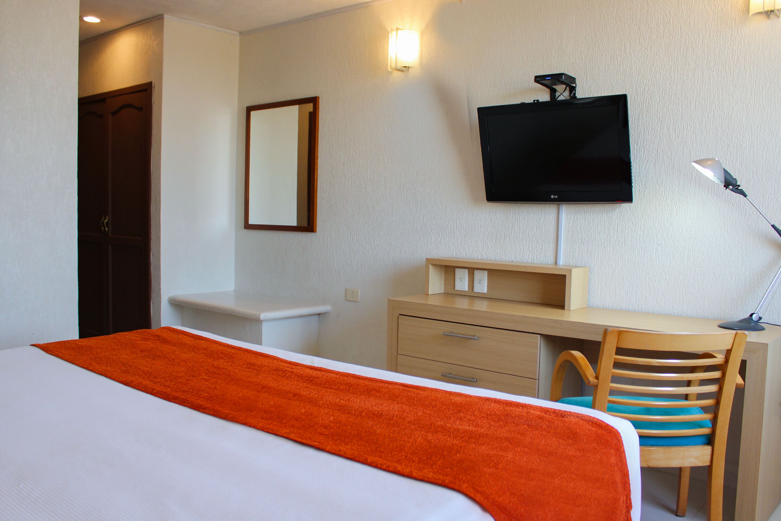 Habitación de hotel con cama, escritorio, silla, televisor y espejo.