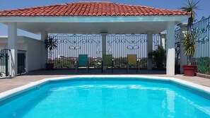 A swimming pool with a tiled roof and metal railing, surrounded by potted plants and outdoor chairs.