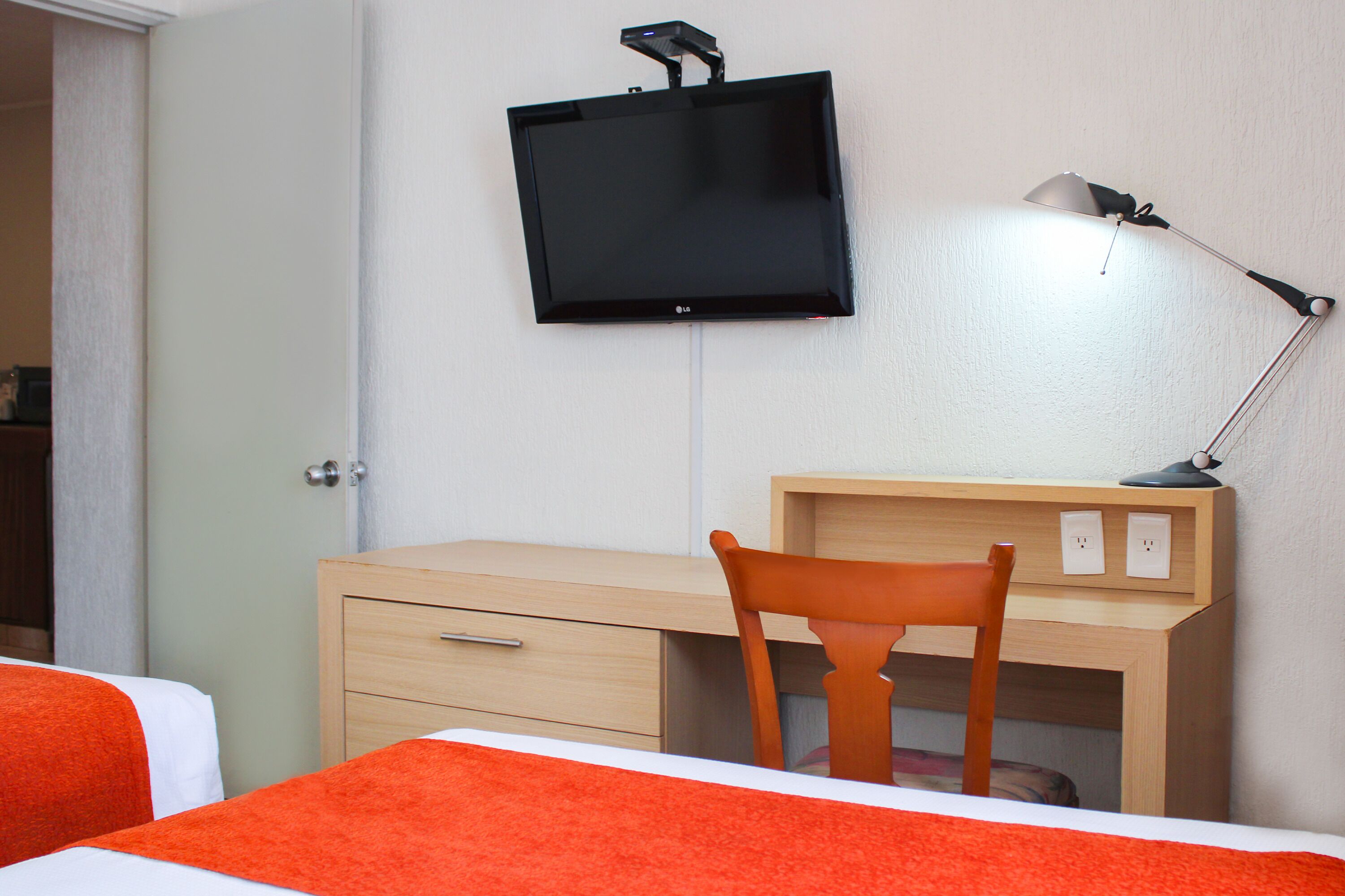 A hotel room with a bed, a desk with a chair, a television mounted on the wall, and a desk lamp.