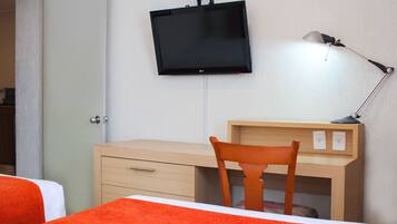 Quarto de hotel com cama, escrivaninha com cadeira, televisão fixada na parede e luminária de mesa.