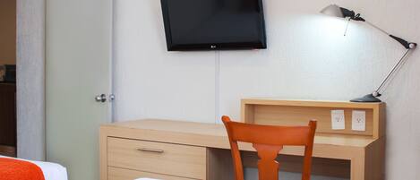 A hotel room with a bed, a desk with a chair, a television mounted on the wall, and a desk lamp.