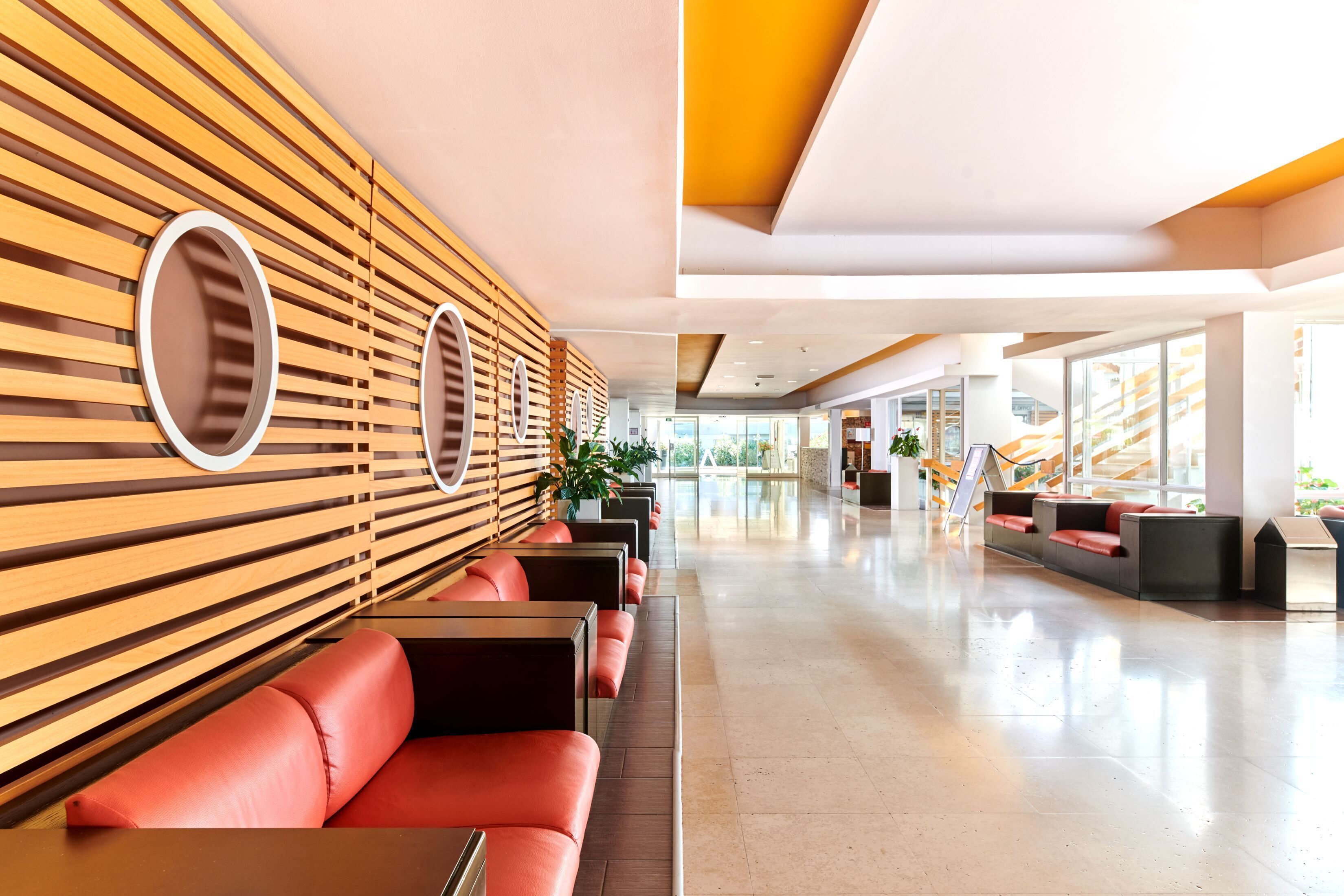 A modern lobby with wooden slats and circular mirrors, red leather seating, and a bright, spacious interior.