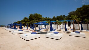 A beachside lounge area with white lounge chairs and blue umbrellas, surrounded by trees and a clear sky. - Hotel Delfin Plava Laguna (Funtana)