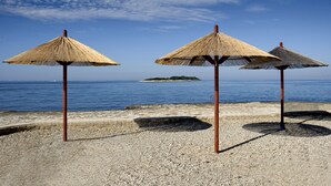 Three straw umbrellas on a pebbled beach with a clear view of the sea and a distant island. - Hotel Delfin Plava Laguna (Funtana)