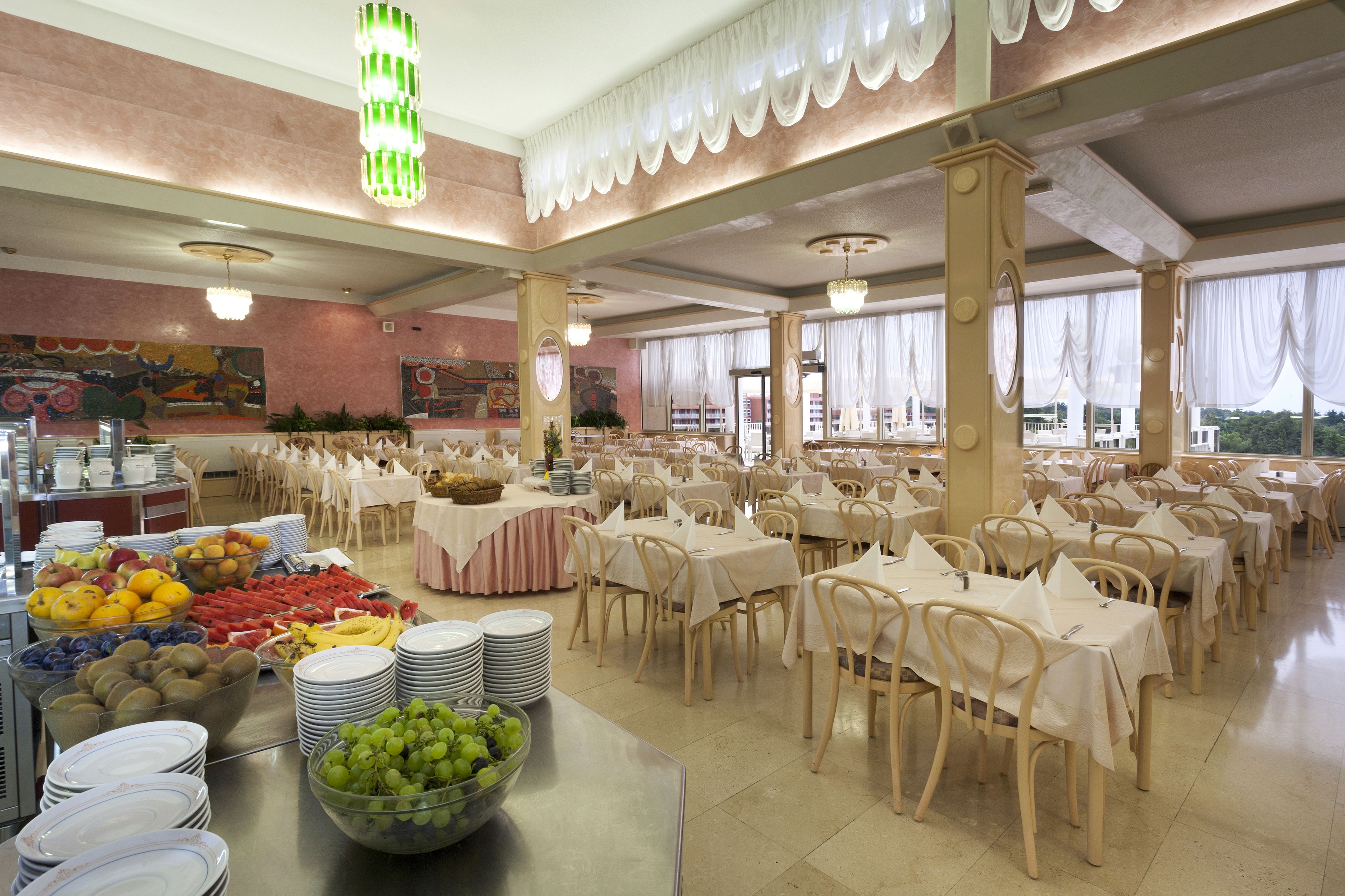Ein geräumiger Essbereich mit mehreren Tischen und Stühlen, ein Buffet mit verschiedenen Früchten und Gemüsen sowie große Fenster mit Vorhängen.