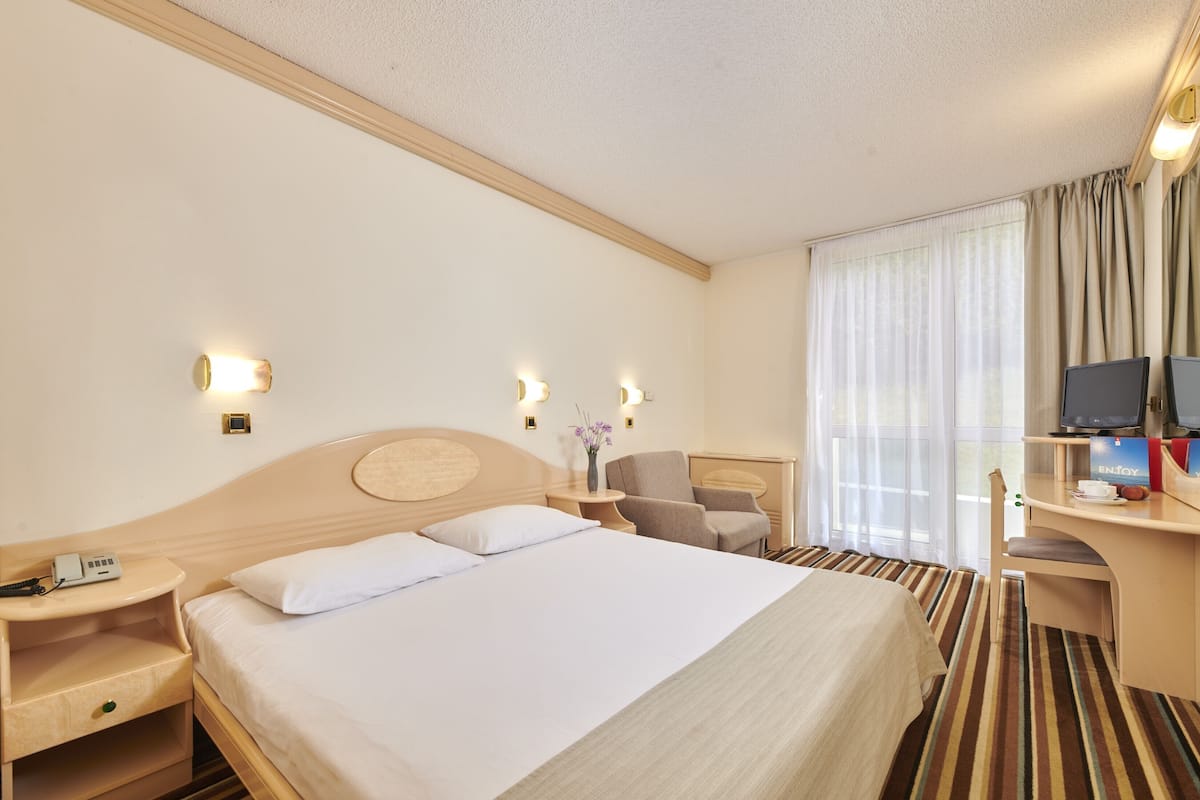 A hotel room with a large bed, bedside tables, a desk with a chair, a television, and a window with curtains.
