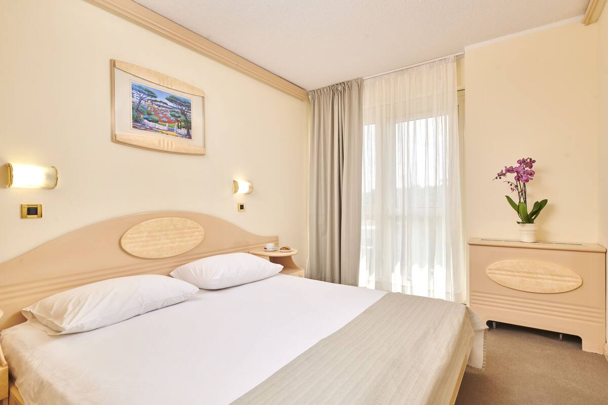 A hotel room with a large bed, a bedside table with a lamp, a small table with a cup and a plate, a painting on the wall, and a vase with flowers on a dresser.