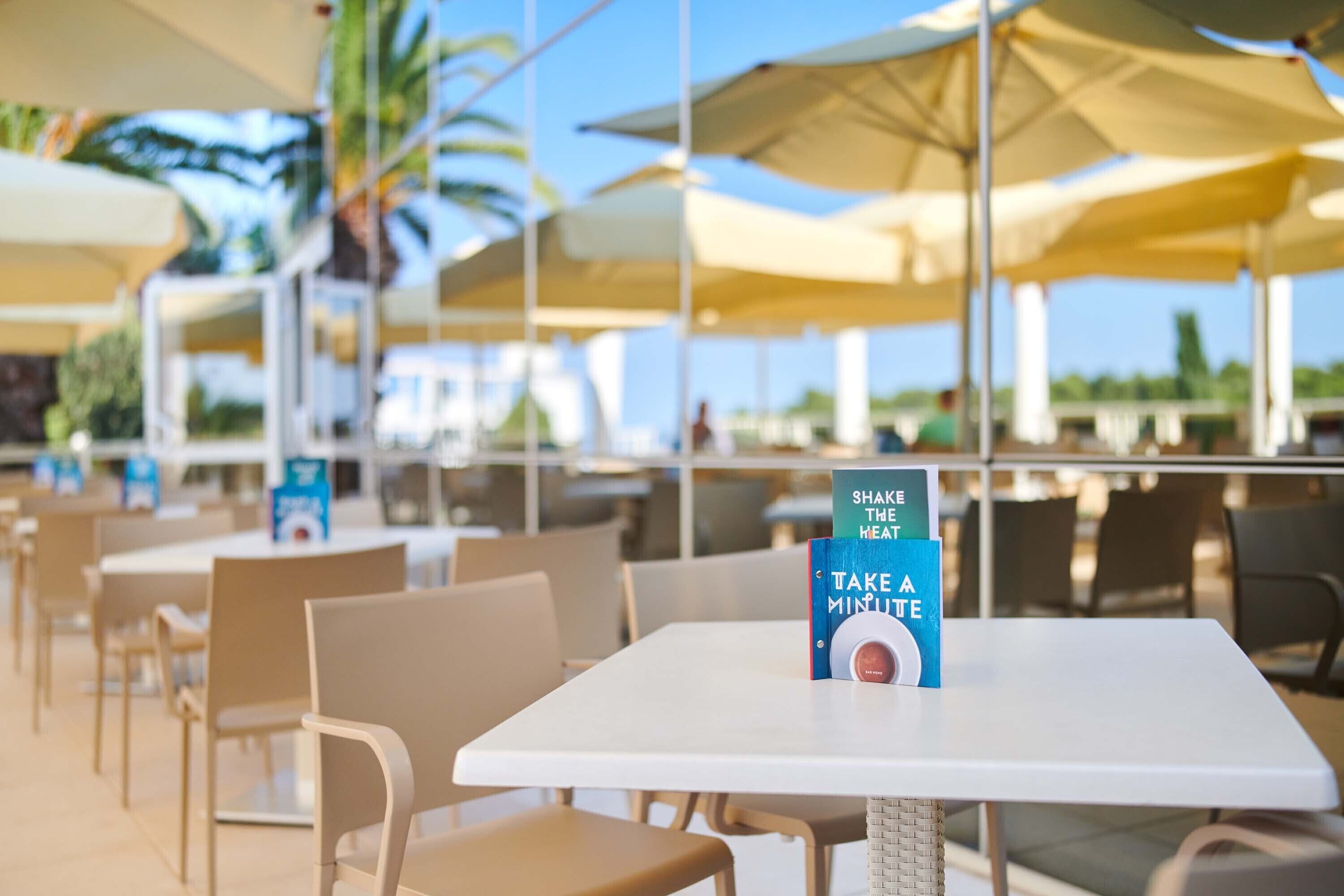 Un área de comedor al aire libre con mesas, sillas, sombrillas y un cartel que promociona una bebida.