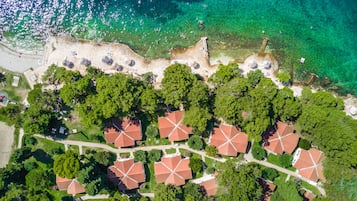 Una vista aerea di una zona costiera con acque turchesi cristalline, una spiaggia sabbiosa e un gruppo di edifici con tetti rossi immersi nel verde.
