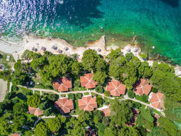An aerial view of a coastal area with clear turquoise waters, a sandy beach, and a cluster of red-roofed buildings surrounded by lush greenery.