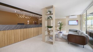 Un espace d’accueil moderne avec un comptoir en marbre, un lambris en bois, une plante en pot, un canapé gris et un écran de télévision.