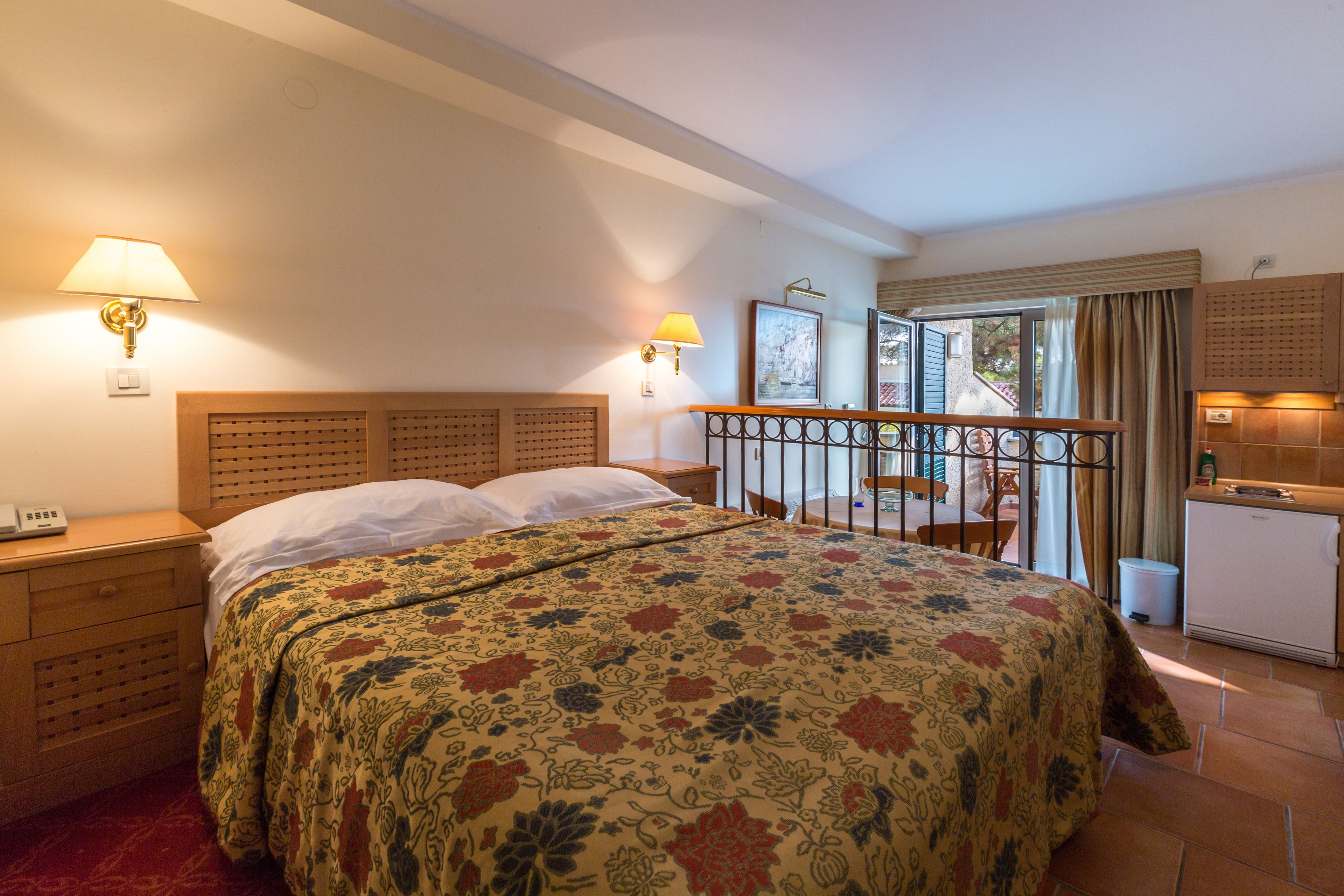 A hotel room with a bed, bedside tables, a desk, a chair, a balcony, and a kitchenette.
