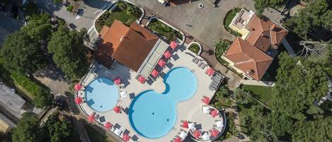 Vue aérienne d’un complexe hôtelier doté d’une grande piscine, de plusieurs parasols rouges et d’un bâtiment central surmonté d’un toit rouge.