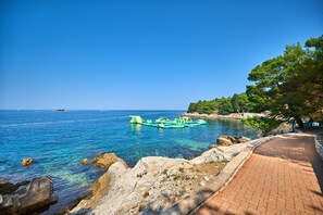 A coastal scene with a clear blue sea, rocky shore, and a paved walkway. - Apartments Bellevue Plava Laguna (Porec)