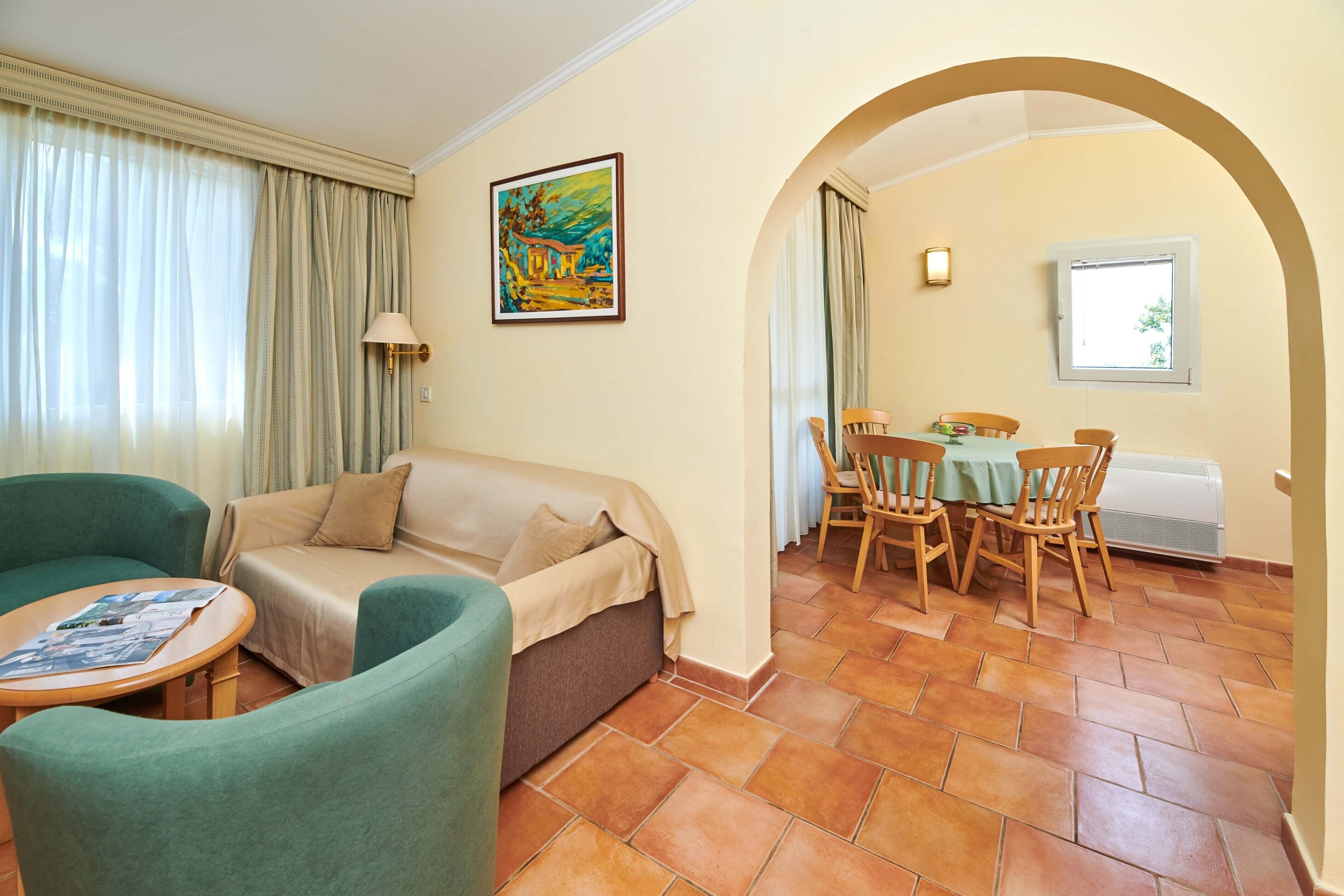 A living room with a sofa, armchairs, a dining table with chairs, a painting on the wall, and a window with curtains.