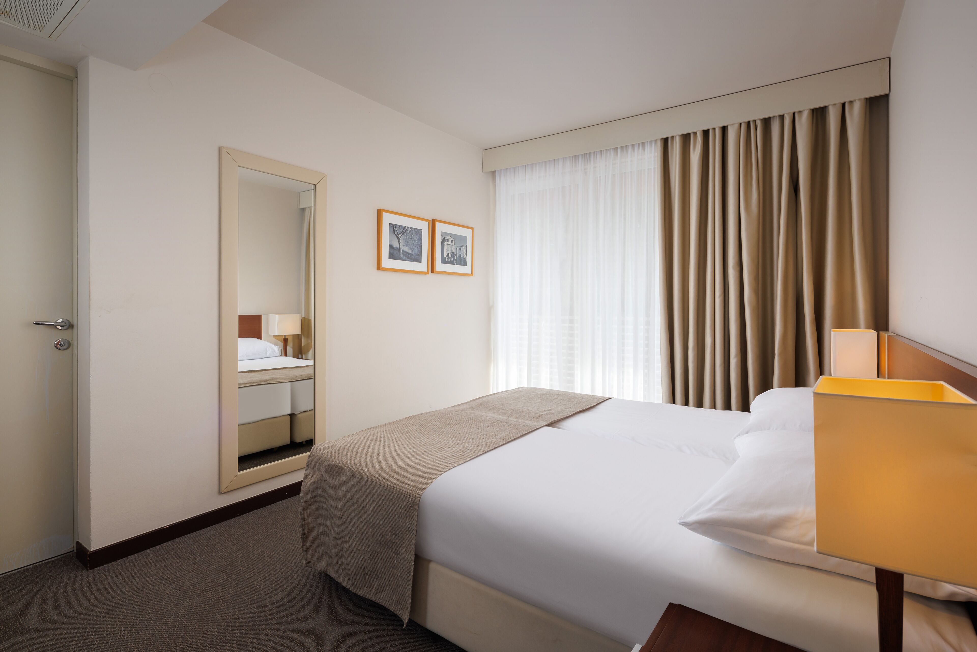 A hotel room with a bed, a bedside lamp, a mirror, a window with curtains, and a door.