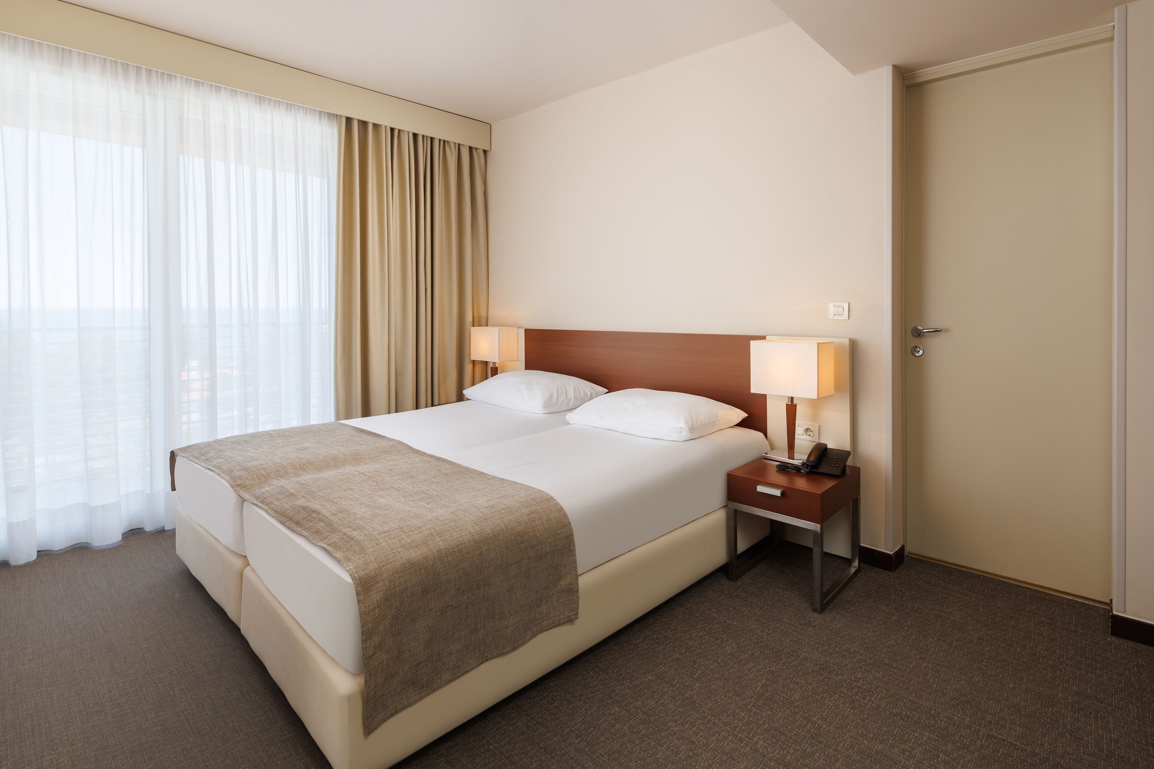 A hotel room with a large bed, two bedside tables with lamps, a door, and a window with curtains.