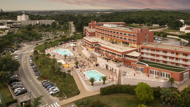 Ein Hotel mit mehreren Schwimmbecken, einem Parkplatz und Blick auf umliegende Grünflächen und Gebäude.
