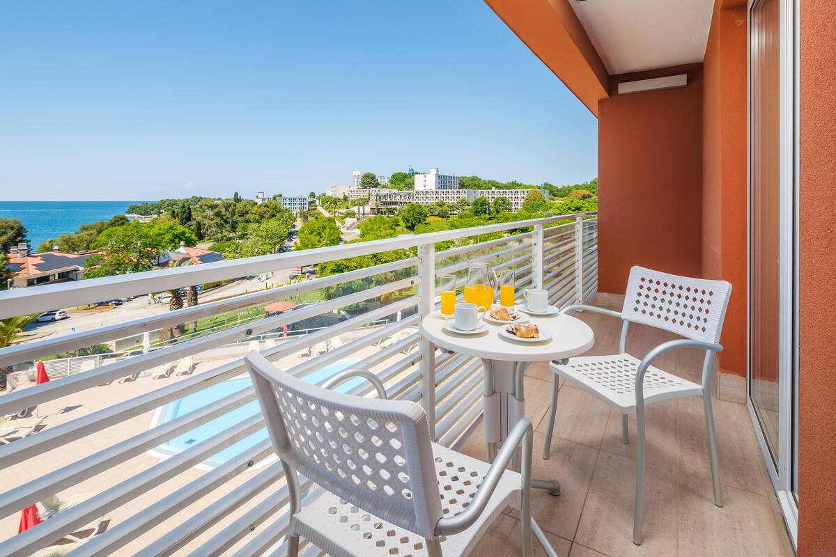 A balcony with white chairs and a table, overlooking a beach and cityscape.