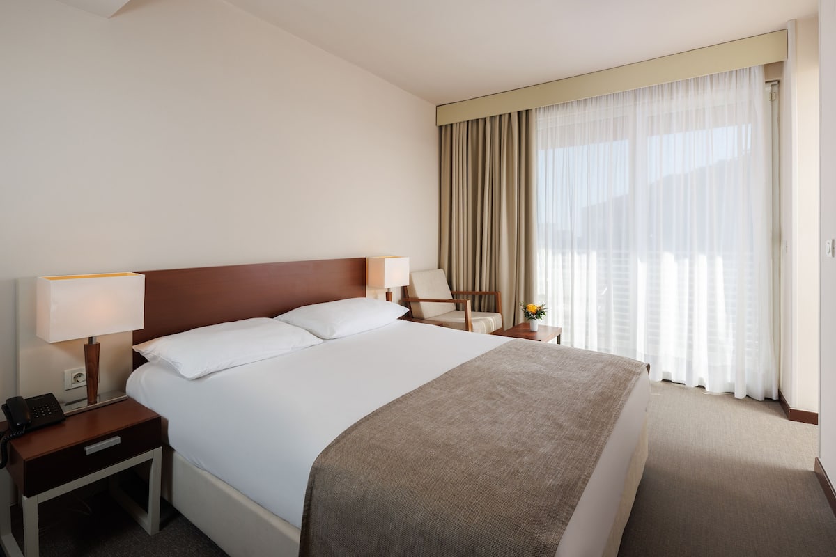 A hotel room with a large bed, two bedside lamps, a nightstand with a phone, a chair, and a window with curtains.