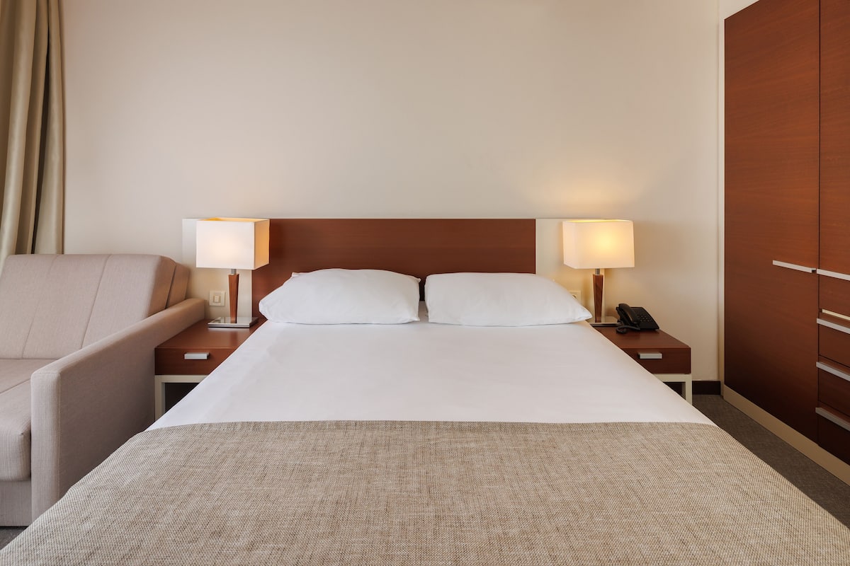 A hotel room with a bed, two bedside tables with lamps, a chair, and a wardrobe.