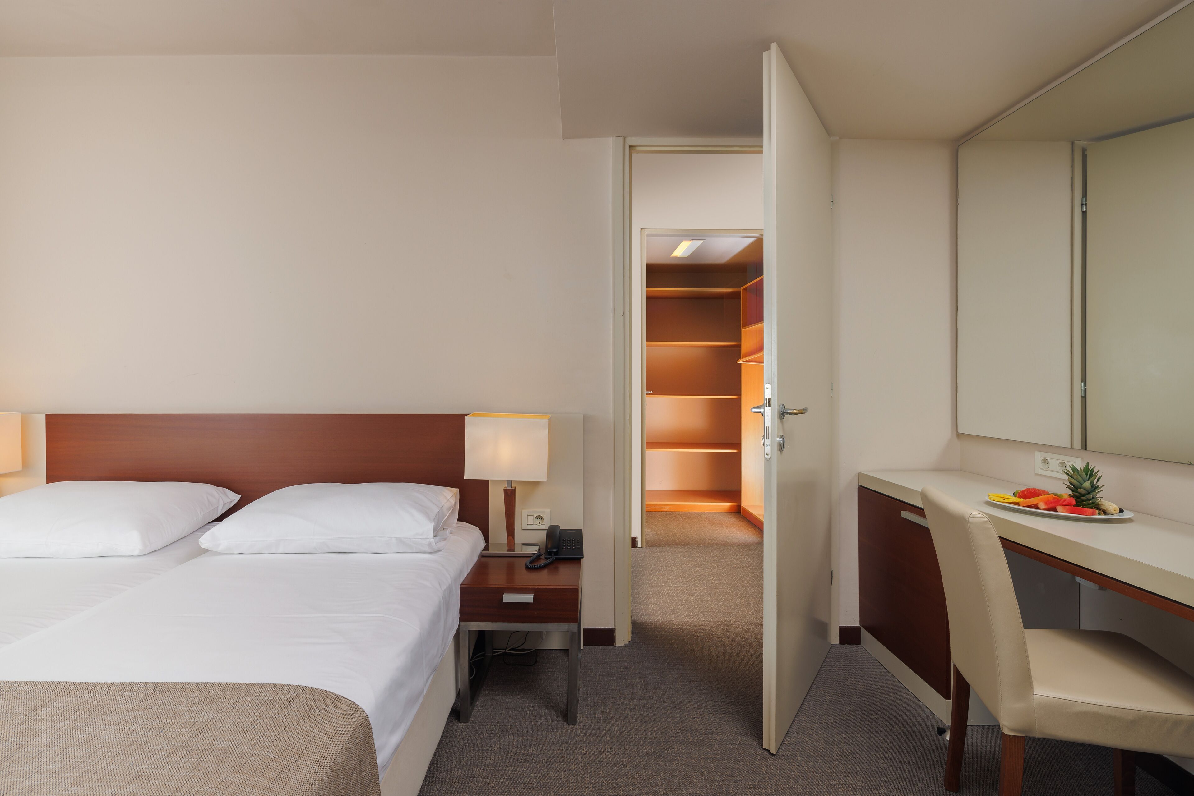A hotel room with a bed, bedside table, lamp, desk, chair, and a small plate with fruit.
