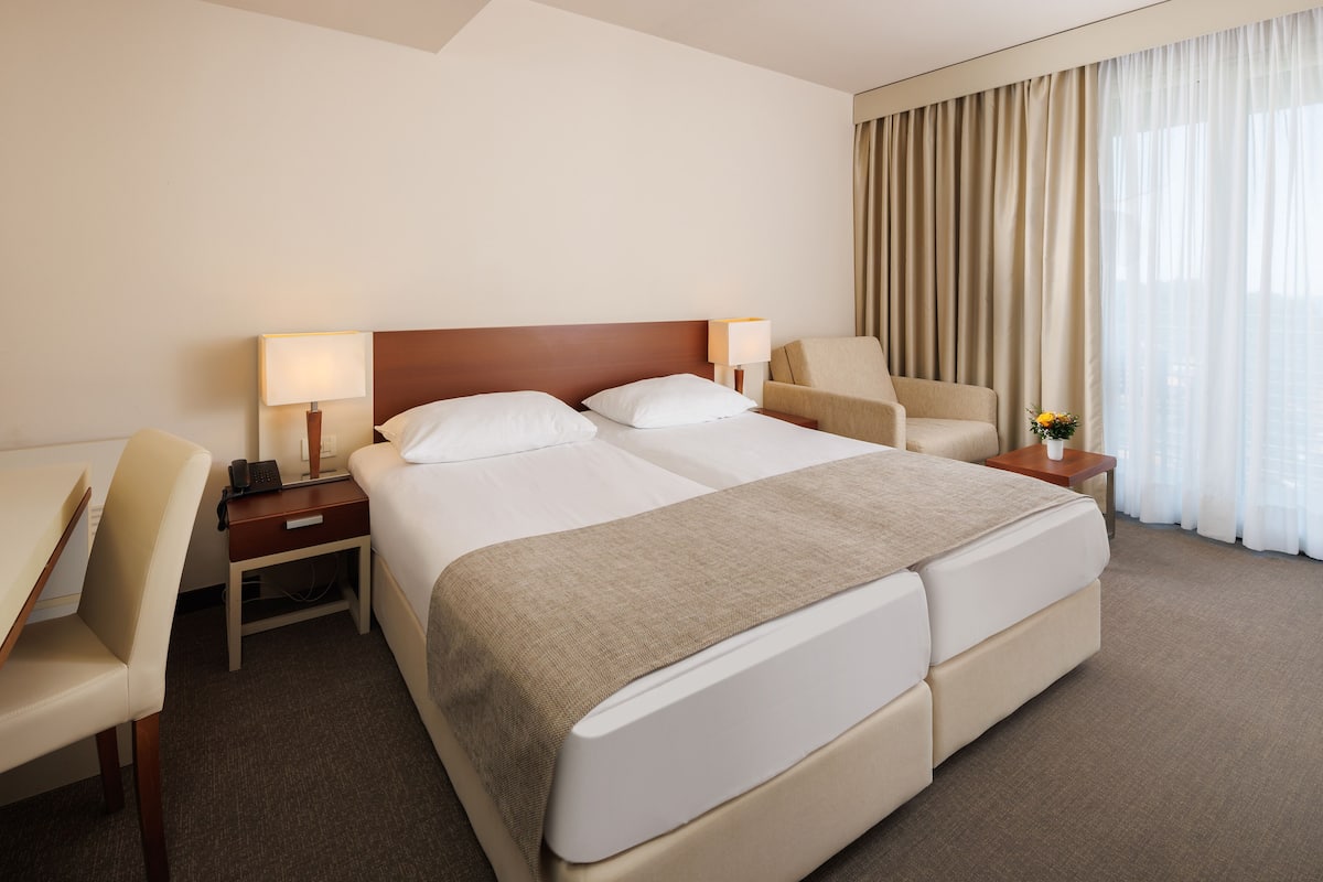 A hotel room with a large bed, a desk, a chair, a lamp, and a view of the city through the window.