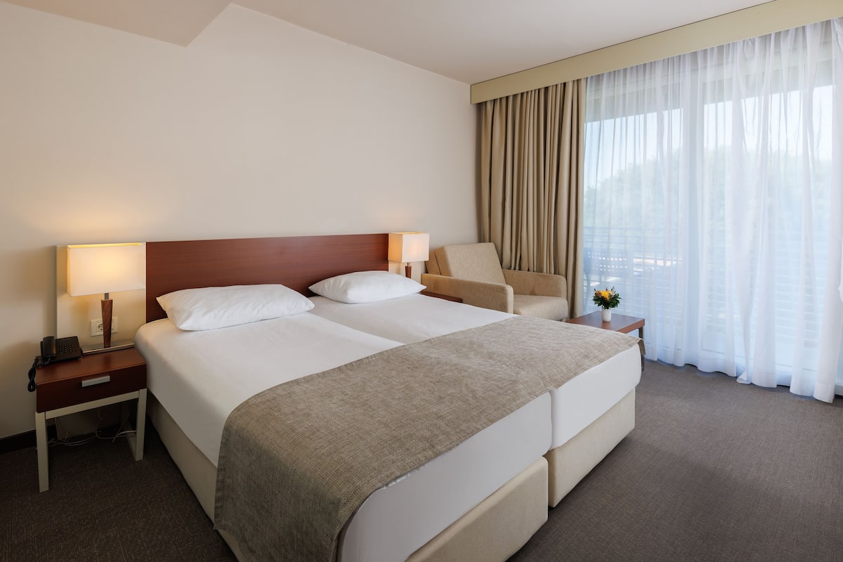 A hotel room with a large bed, a chair, a nightstand with a lamp, and a window with curtains.