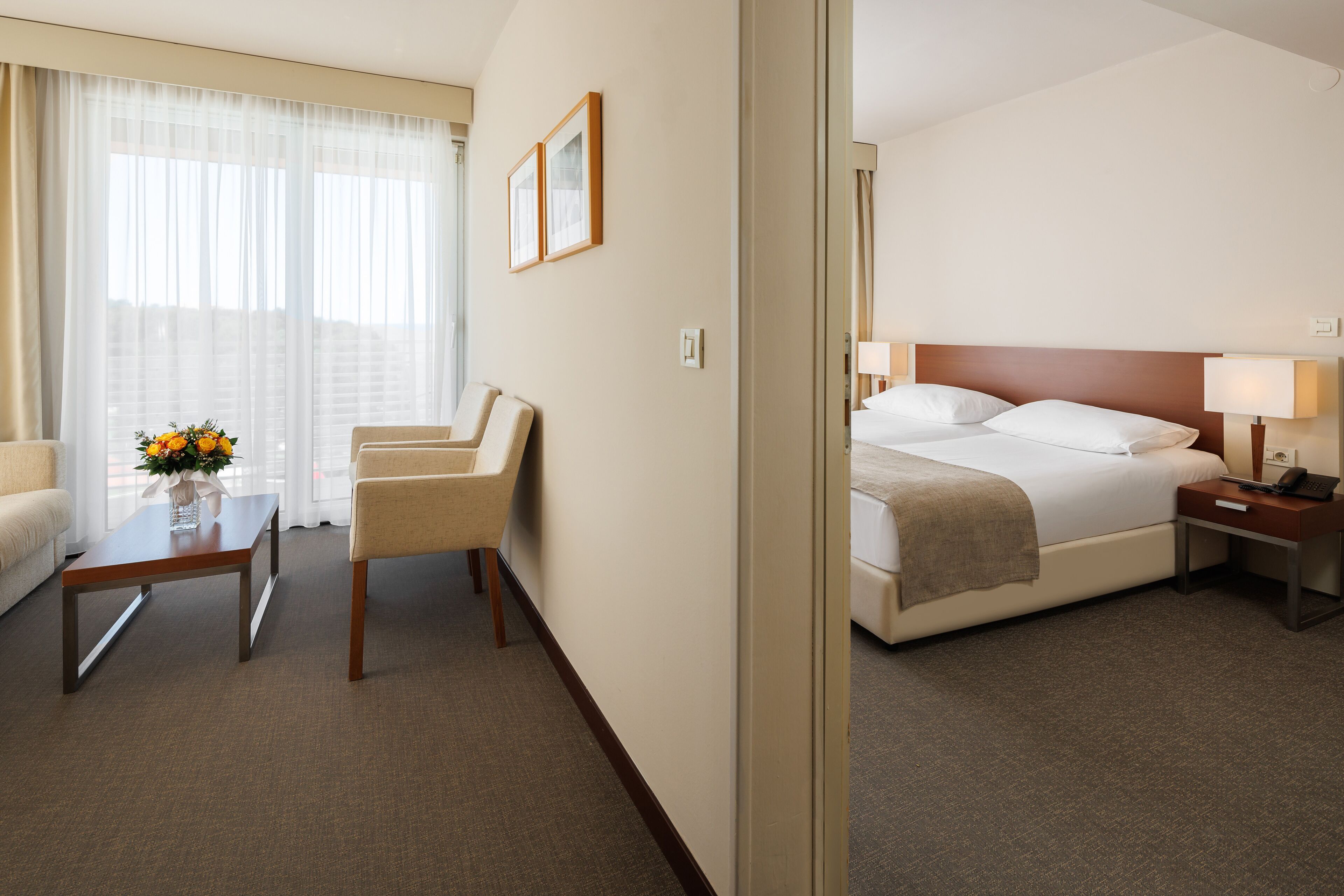 A hotel room with a bed, a desk, a chair, a small table with a vase of flowers, and a window with sheer curtains.