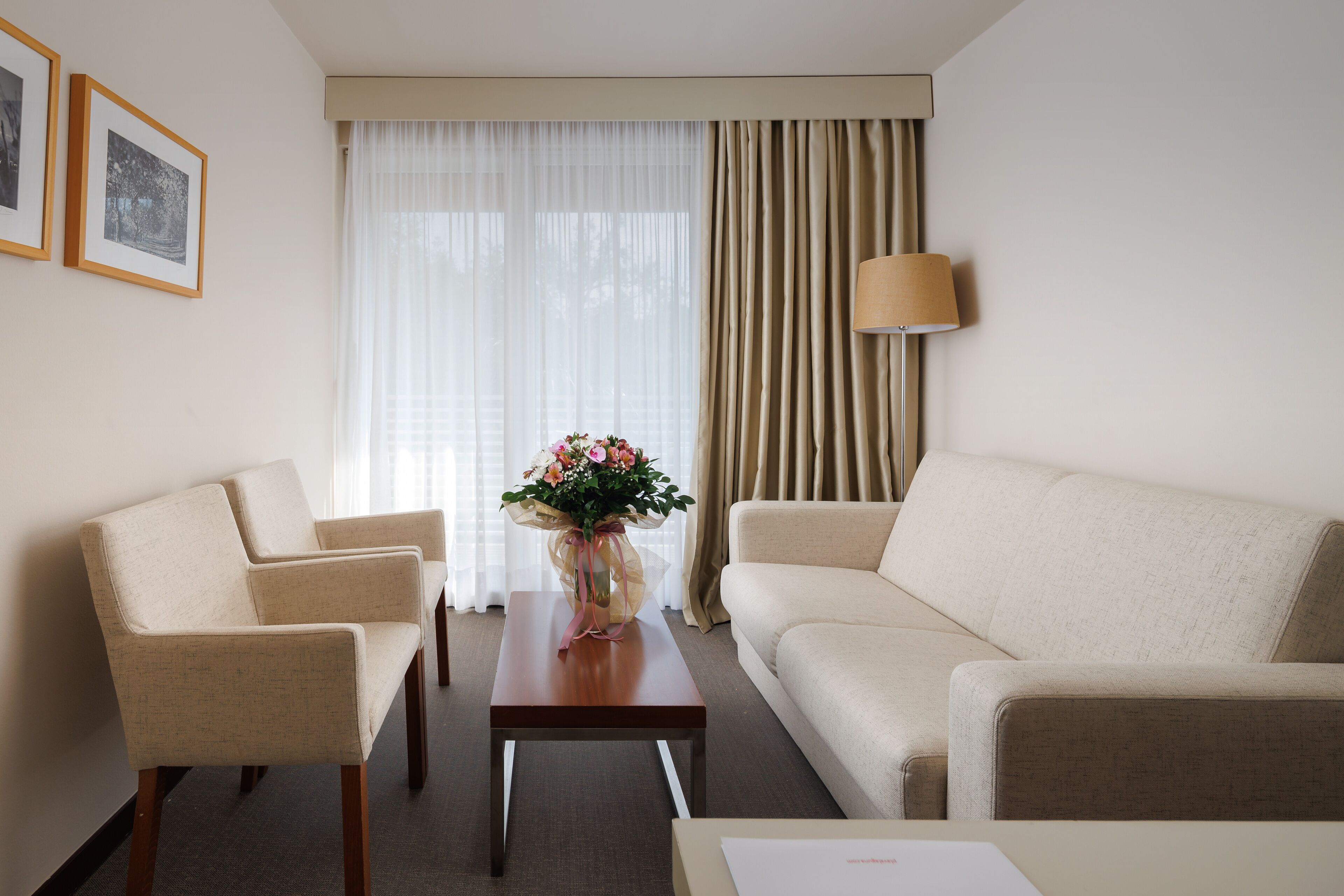 Een hotelkamer met een beige bank, twee fauteuils, een houten salontafel met een bloevas, een lamp en een groot raam met dunne gordijnen.