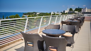 Eine Terrasse mit Korbstühlen und Tischen mit Blick auf eine Küstenlandschaft, einschließlich Gebäude und Grünflächen.