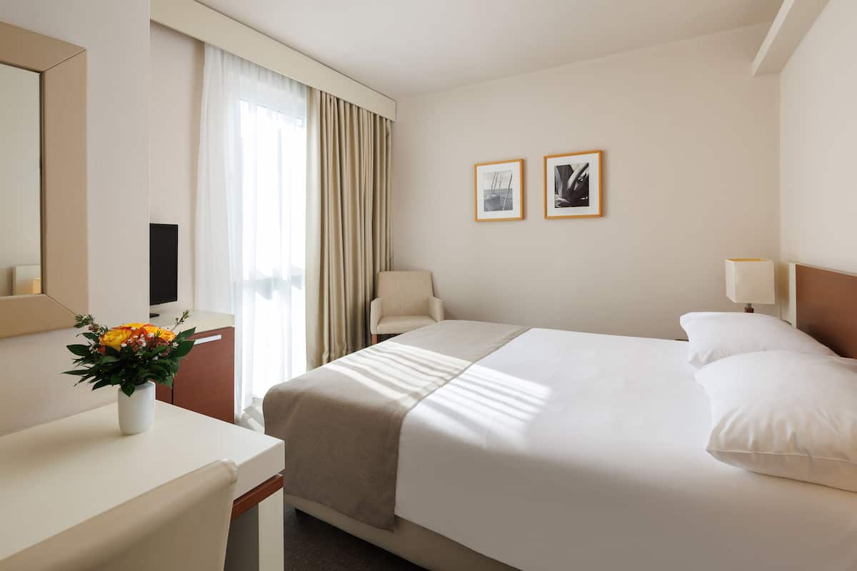 A hotel room with a bed, a desk, a chair, a TV, a window with curtains, and two framed pictures on the wall.