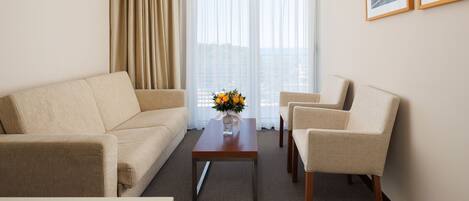 Quarto de hotel com sofá bege, poltrona combinando, mesa de centro de madeira com um vaso de flores, e uma janela grande com cortinas leves.