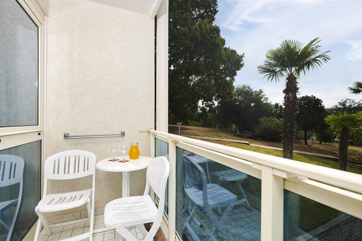 Een balkon met witte plastic stoelen en een kleine tafel, een glazen hekwerk en uitzicht op bomen en een palmboom.