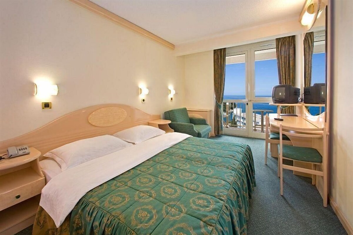 A hotel room with a bed, a desk with a chair, a television, and a view of the sea through the window.