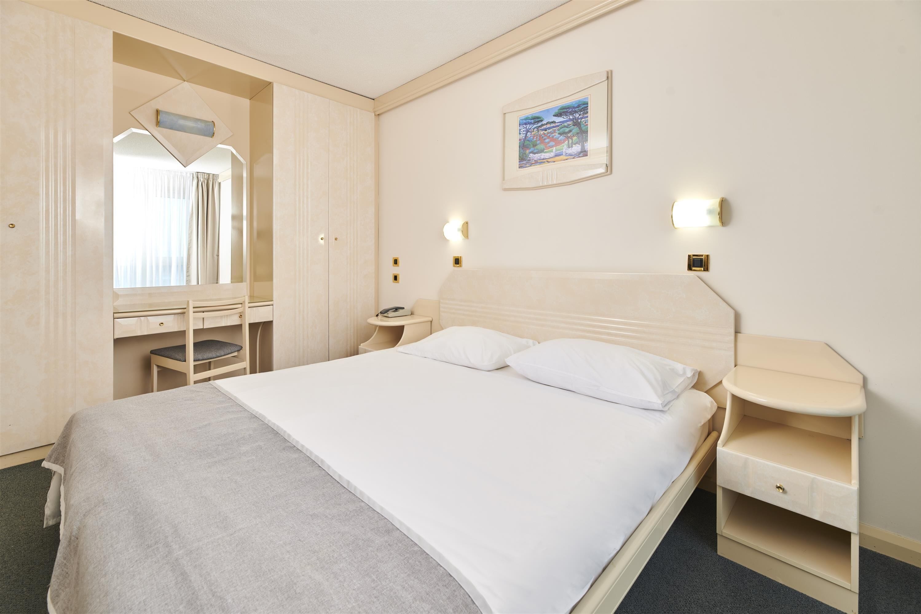 Una camera d'albergo ordinata con un letto, comodini, una scrivania con una sedia, un armadio e un quadro appeso al muro.
