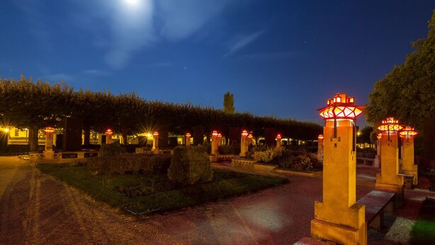 A nighttime scene with a paved pathway, illuminated by street lamps, leading through a landscaped area with trimmed hedges and benches. - Arche Hotel (Vierzon)
