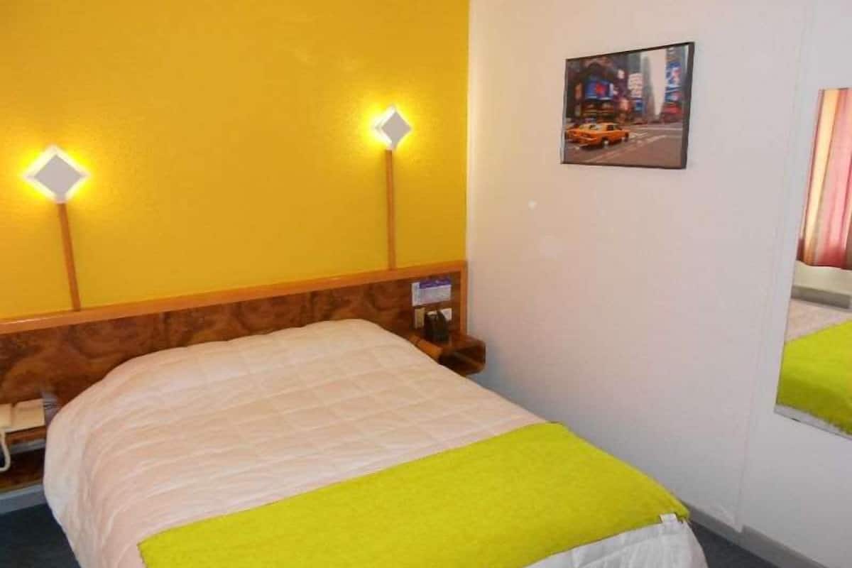 A hotel room with a bed, a nightstand, a wall-mounted lamp, and a framed picture on the wall.