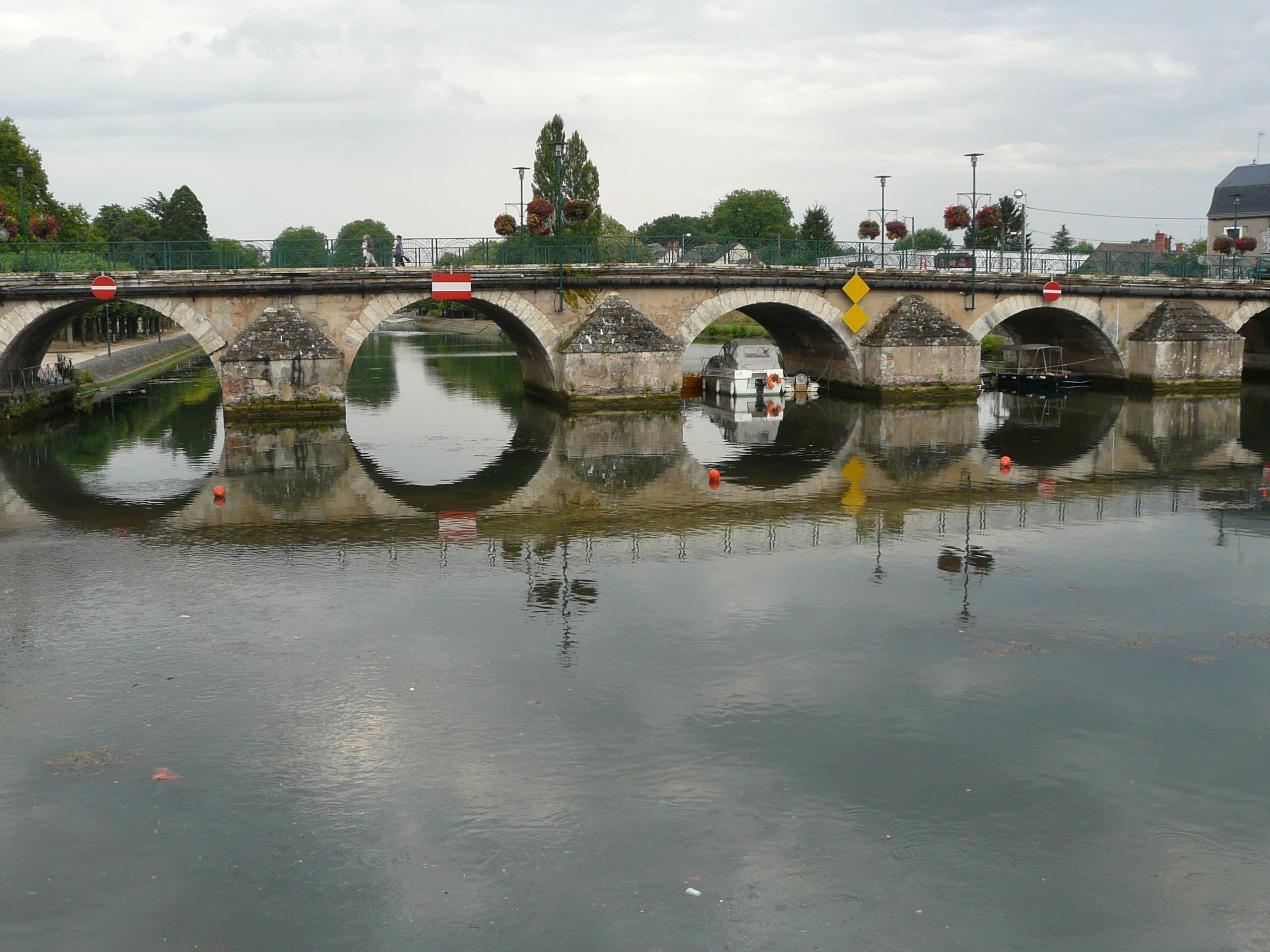 A stone bridge over a calm river with a reflection of the bridge and surrounding greenery.