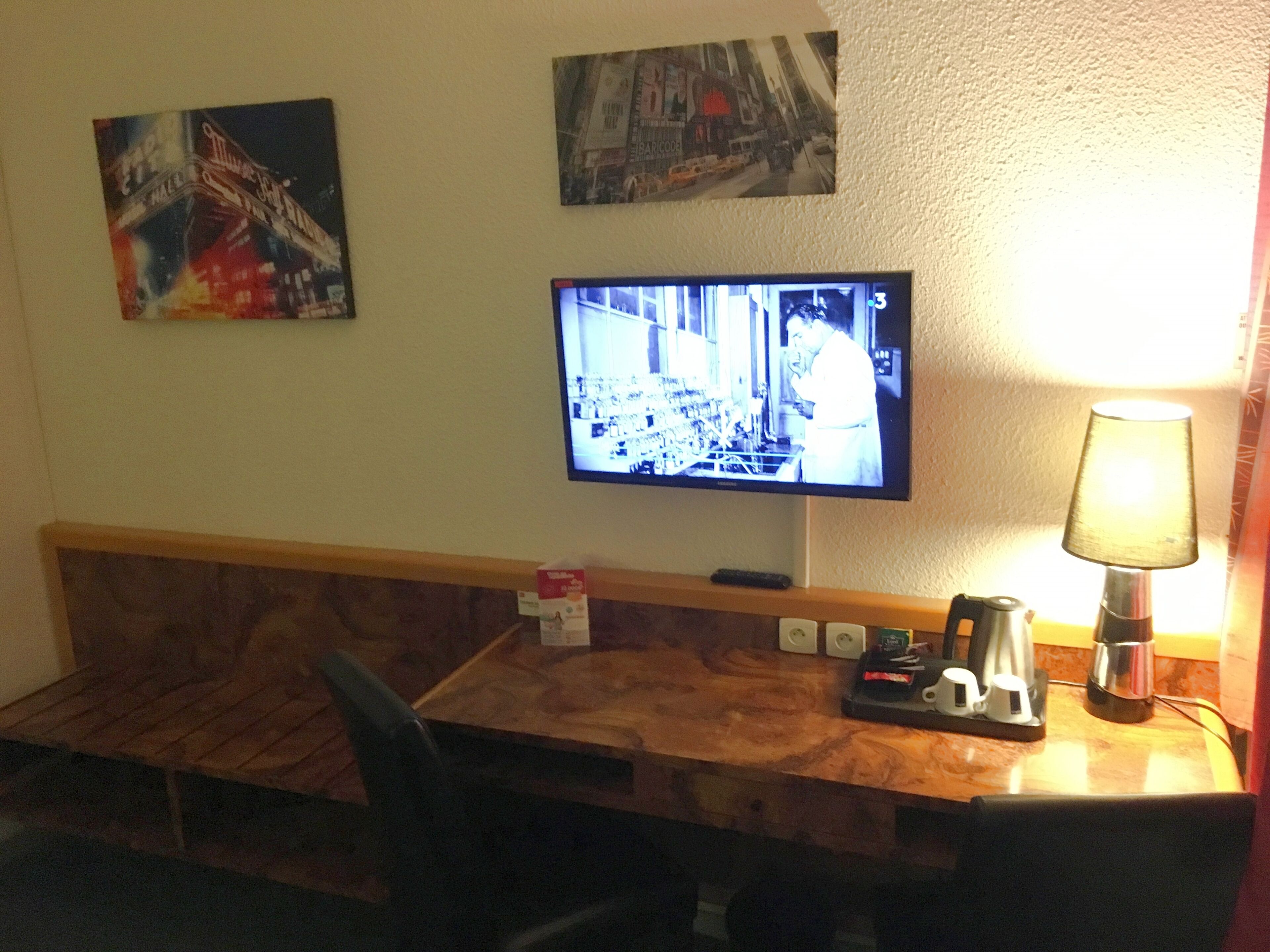 A hotel room with a wooden headboard, a television mounted on the wall, a lamp, and a small table with a coffee maker and cups.