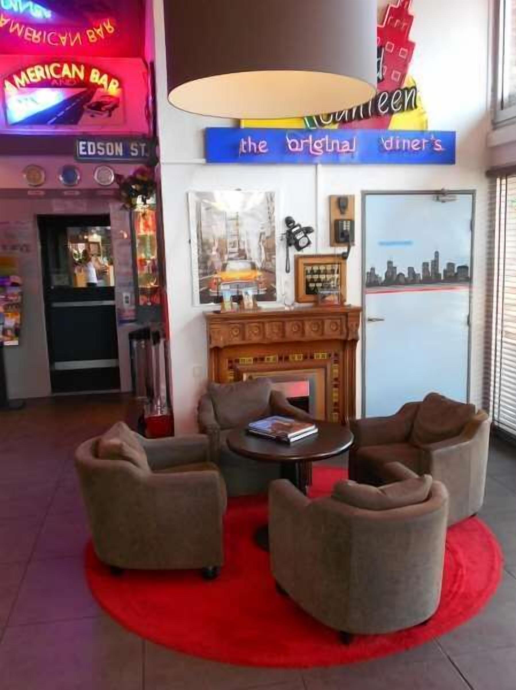 A lobby with a neon sign for 'American Bar' and 'The Original Diner's'. There are four armchairs around a small table, a fireplace, and a cityscape view through the door.